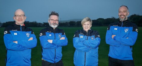 Four individuals standing outside with arms crossed, wearing blue and black jackets with team logos, and smiling for the camera. A grassy field is in the background.