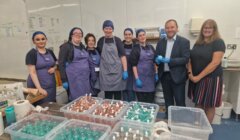 A group of people, some in aprons and hair nets, pose in front of a table filled with containers of variously colored liquids in a laboratory or production setting.