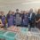 A group of people, some in aprons and hair nets, pose in front of a table filled with containers of variously colored liquids in a laboratory or production setting.