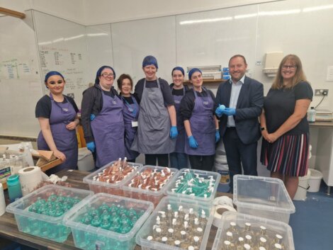 A group of people, some in aprons and hair nets, pose in front of a table filled with containers of variously colored liquids in a laboratory or production setting.