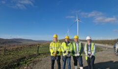 Four individuals wearing high-visibility clothing and hard hats stand on a gravel path near wind turbines in a hilly area.