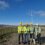 Four individuals wearing high-visibility clothing and hard hats stand on a gravel path near wind turbines in a hilly area.