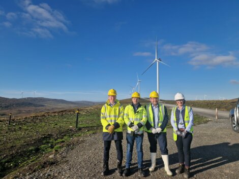 Four individuals wearing high-visibility clothing and hard hats stand on a gravel path near wind turbines in a hilly area.