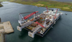 A large offshore construction vessel with cranes docked in a calm body of water near a hilly landscape.