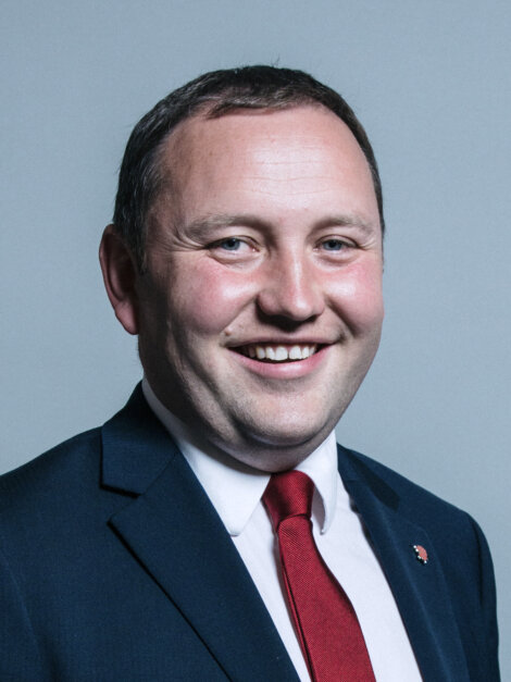 A man in a suit and red tie smiles at the camera against a plain background.