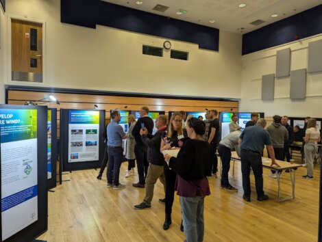 A group of people are gathered in a room with wooden floors and display boards presenting information. Some individuals are conversing, and others are reading the displays.