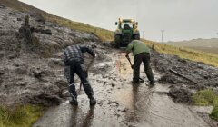 Two individuals are clearing mud and debris from a rural road with shovels as a tractor assists in the background. The scene takes place on a wet, overcast day.