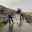 Two individuals are clearing mud and debris from a rural road with shovels as a tractor assists in the background. The scene takes place on a wet, overcast day.