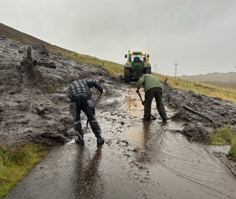 Two individuals are clearing mud and debris from a rural road with shovels as a tractor assists in the background. The scene takes place on a wet, overcast day.