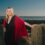 A woman with long blonde hair, wearing a black dress and red shawl, stands on a rocky coastline with her hand on a cement structure, with the sea and a clear sky in the background.