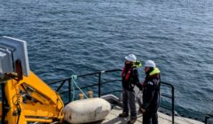 Two people in safety gear stand on a boat's deck, monitoring equipment. An island is seen in the distance across the water.