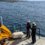 Two people in safety gear stand on a boat's deck, monitoring equipment. An island is seen in the distance across the water.