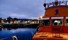 An orange RNLI lifeboat docked at a harbor with town buildings and evening lights in the background under a cloudy sky.