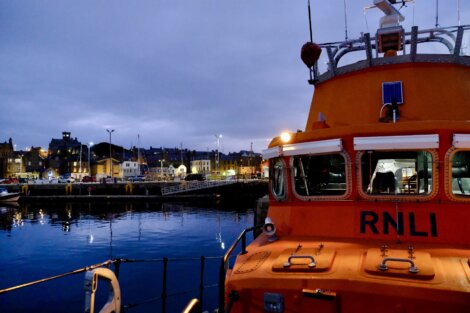 An orange RNLI lifeboat docked at a harbor with town buildings and evening lights in the background under a cloudy sky.