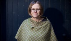 A woman with short hair and glasses is standing in front of a dark background, wearing a green polka dot patterned wrap.