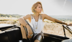 A person with short hair sits in a convertible car, looking into the distance. They are wearing a white tank top and shorts. The background features a sunny landscape with hills and vegetation.