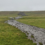 A muddy, meandering path cuts through a green grassy field on a cloudy day, with a small hill visible in the background.