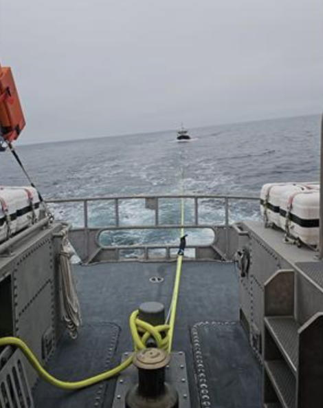 A boat is being towed behind another vessel across a vast, calm ocean with a cloudy sky. The deck of the towing vessel is visible with safety equipment on both sides and a towline secured in the middle.