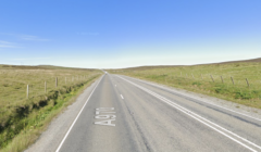 A straight stretch of the A970 road in a rural area with grass fields on both sides, under a clear blue sky.