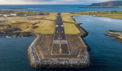 Aerial view of a coastal runway extending into the sea with adjacent green fields and small buildings.