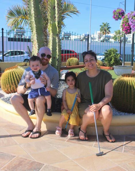 A family of four, including two children and two adults, poses for a photo next to a cactus in a sunny outdoor mini-golf area. The adults are seated, holding mini-golf clubs.