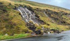 A hillside with visible erosion shows streams of water and mud flowing down towards a roadside.