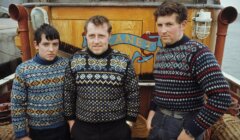 Three men in patterned sweaters pose in front of a wooden boat named "Clyne.