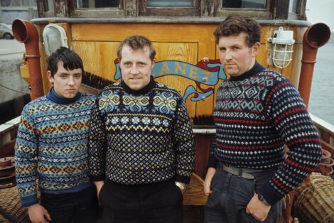 Three men in patterned sweaters pose in front of a wooden boat named "Clyne.