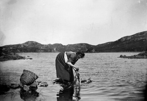 A person in a dress is bending over to wash clothes in a lake, with a wicker basket on the ground beside them and mountains in the background.