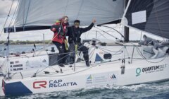 Two sailors on a yacht celebrate during a regatta. The yacht is branded with various logos and the text "Quantum Sails," with a shoreline in the background.