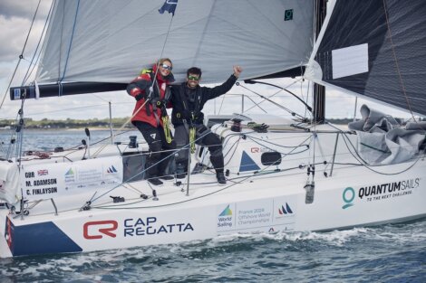 Two sailors on a yacht celebrate during a regatta. The yacht is branded with various logos and the text "Quantum Sails," with a shoreline in the background.