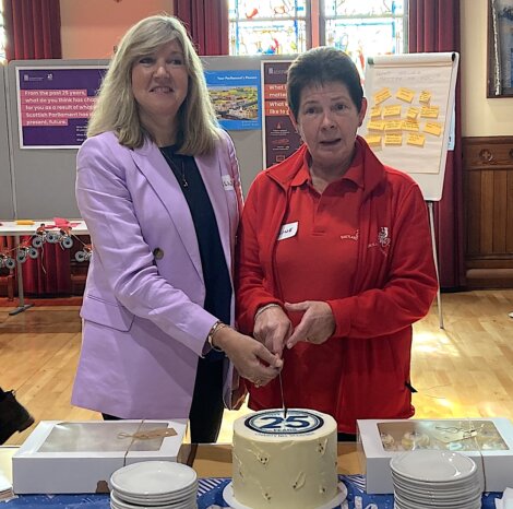 Two people cutting a cake with "25" on it. One is wearing a light purple blazer, and the other is in a red sweater. Display boards and a flip chart are in the background.