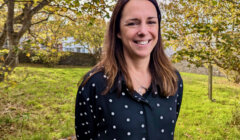 Woman with long hair wearing a black polka-dot shirt stands outside on a grassy area with trees in the background, smiling at the camera.