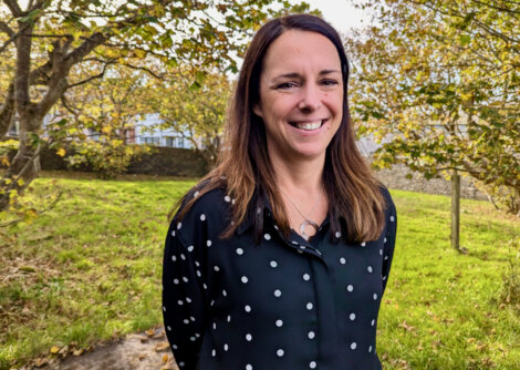 Woman with long hair wearing a black polka-dot shirt stands outside on a grassy area with trees in the background, smiling at the camera.