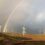 Wind turbines on a grassy landscape with a rainbow in the cloudy sky.