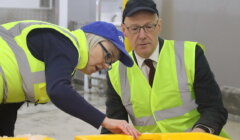 Two people wearing high-visibility vests and caps examine bins filled with ice cubes, inside an industrial setting.