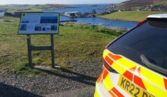 A police car is parked on a scenic overlook with a view of a coastal town and body of water. An informational sign is mounted nearby.