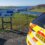 A police car is parked on a scenic overlook with a view of a coastal town and body of water. An informational sign is mounted nearby.