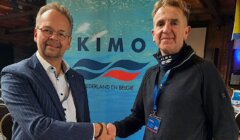 Two men shaking hands in front of a "KIMO Nederland en België" banner, one wearing a striped suit and the other in a black jacket.