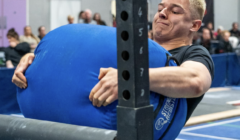 Person lifting a large blue sandbag during a competition, with spectators in the background.