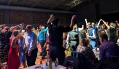 A group of people dancing energetically indoors, with others seated at tables front right. The room is dimly lit with a spacious ceiling.