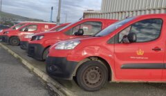 Four red Royal Mail vans parked in a row at a lot, with a cloudy sky and industrial buildings in the background.