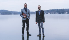 Two men stand on an icy surface; one holds a violin. Trees and water are visible in the background.