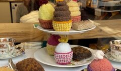 Display of knitted cupcakes on a tiered stand with teacups and saucers on the table, viewed through a window with a man and parked car in the background.