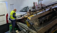 A worker in a yellow vest examines a damaged car and machinery on a ferry. The car is partially crushed, and there are visible vehicle parts and cables.