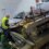 A worker in a yellow vest examines a damaged car and machinery on a ferry. The car is partially crushed, and there are visible vehicle parts and cables.