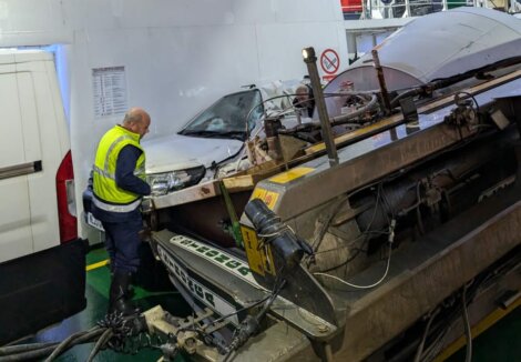 A worker in a yellow vest examines a damaged car and machinery on a ferry. The car is partially crushed, and there are visible vehicle parts and cables.