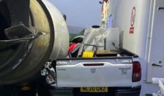 A white pickup truck with the license plate NL20 GNZ is damaged and crushed between a ferry wall and a large cylindrical object.