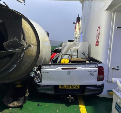 A white pickup truck with the license plate NL20 GNZ is damaged and crushed between a ferry wall and a large cylindrical object.