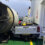 A white pickup truck is crushed under a large industrial mixer on a ferry deck. The truck bed is exposed, and a no-smoking sign is visible on the wall.